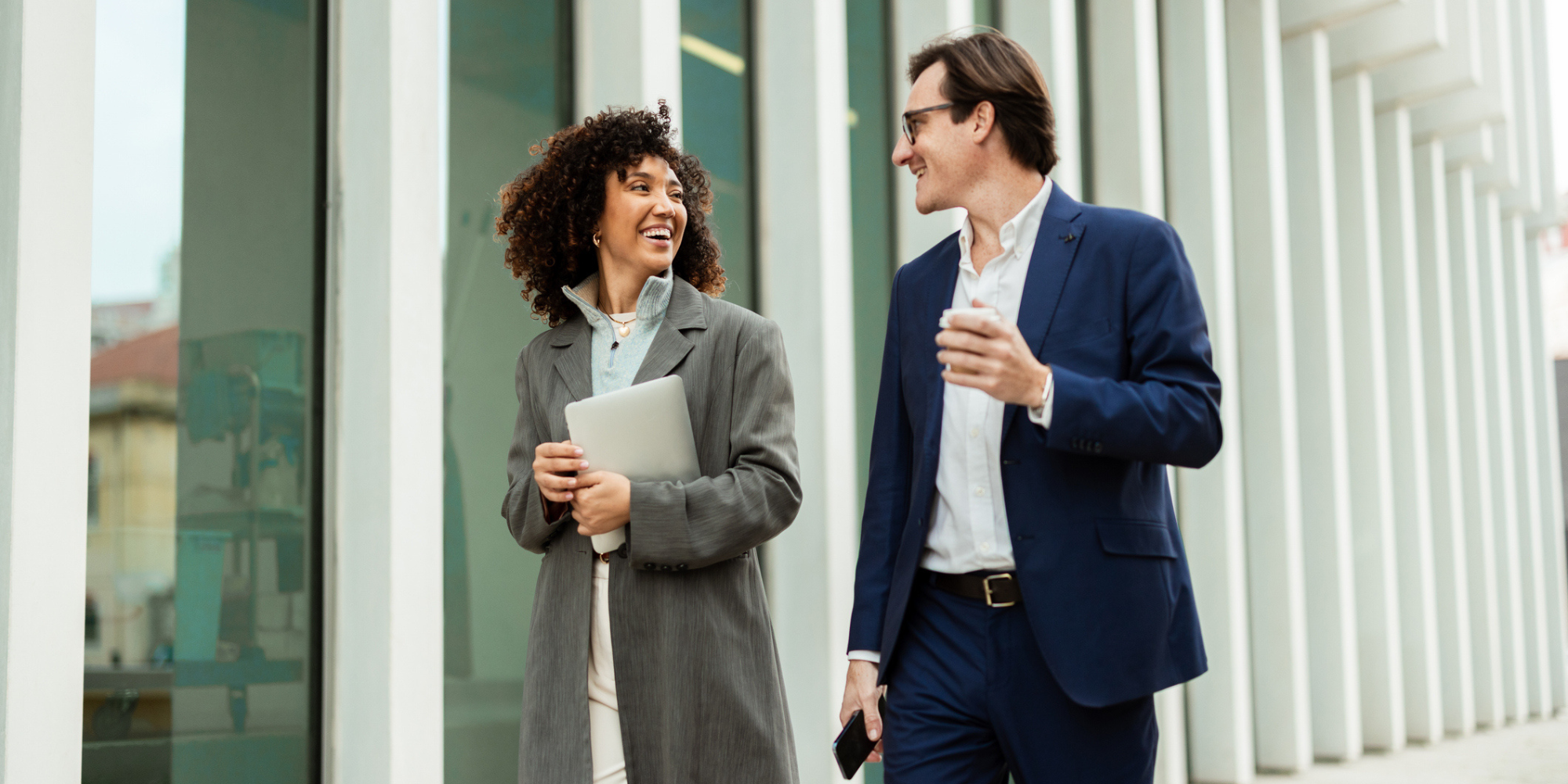 A woman and a man walk together, smiling, outside a modern building, engaged in conversation.